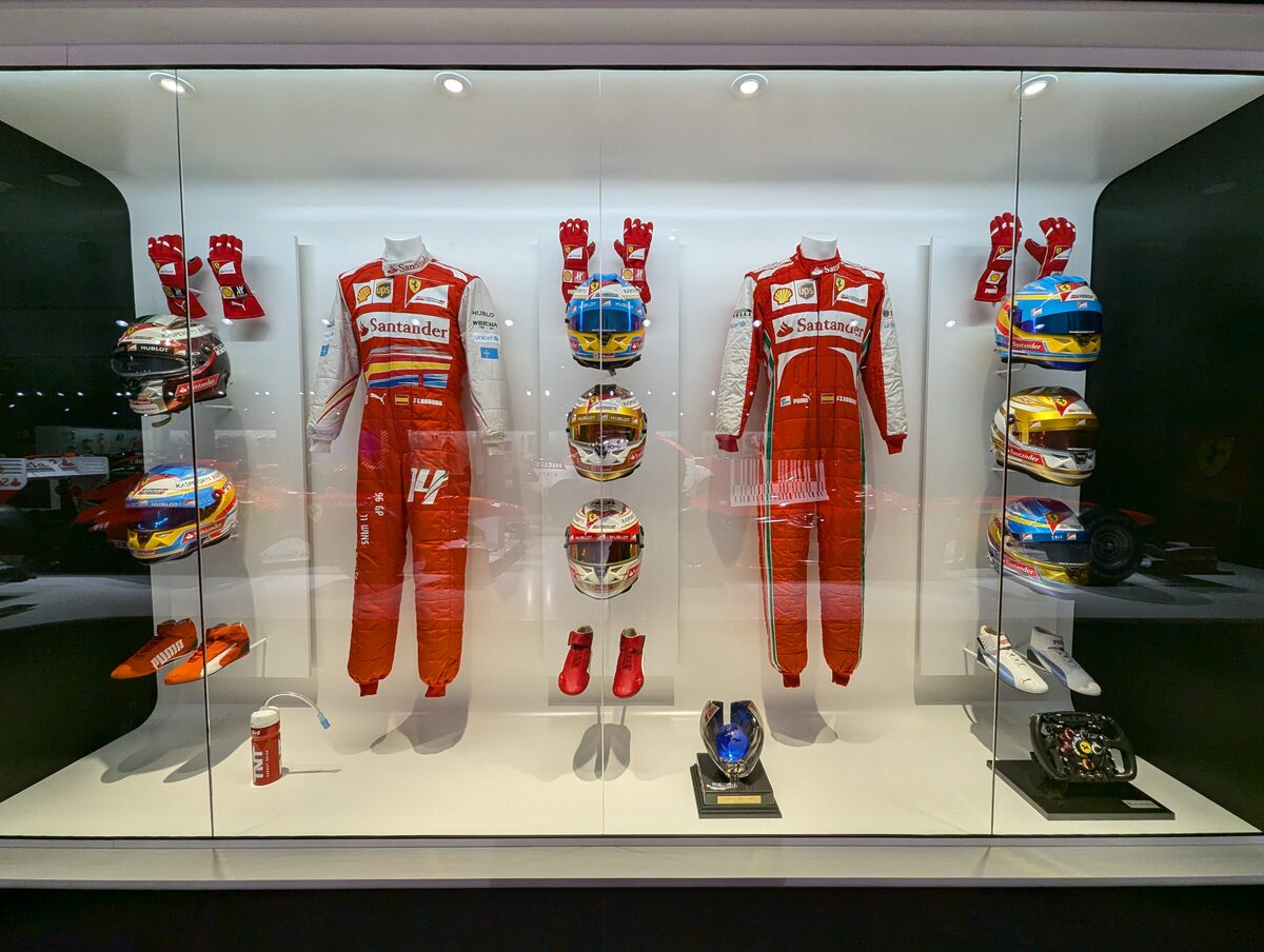 フェラーリ時代のレーシングスーツ2着とヘルメット・グローブ・シューズ・ステアリングの展示ケース